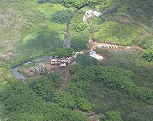 An overhead view of a set of buildings located in a dense jungle. A river runs to the left of the buildings, and moving trucks are seen to the right of the buildings. There is a dirt road leading from the buildings to other buildings.