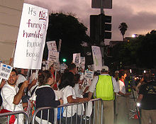 A crowd of people at the left are standing behind a gated barrier. The crowd is holding protest signs and the majority are looking away from the camera. An obscured man is being interviewed at the right side of the image. The sky appears to be approaching night.