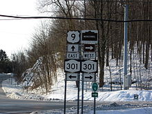 A series of road signs in front of an intersection between two highways. The hill behind the intersection is covered in snow; electrical wires hang overhead.