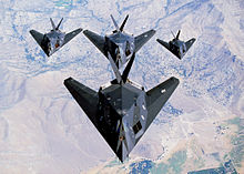 Look-down view of four black aircraft flying in formation, three of which fly behind and underneath the lead aircraft.
