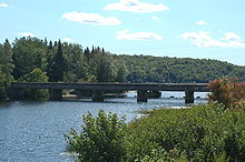 A river flowing between two banks covered in vegetation and trees. The river flows under a concrete bridge resting on four support piers into a lake in the background
