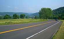 A paved two-lane road crosses the lower half of the picture from left to right. A small blue car is visible at the right with its brake lamps on, where the road turns back sharply. After the turn it is slightly downhill from the camera and cannot be seen, although black-on-yellow arrow signs indicate where it is. In the distance, in the top of the image, is a valley with green hills on either side