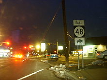 A nighttime view of a road lined with businesses approaching a traffic light. A sign on the right side of the road reads east Route 48 right.