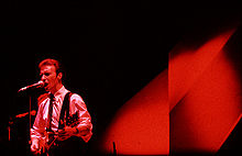 A colour photograph of members of Midge Ure of the band Ultravox performing on a stage with a microphone and a guitar
