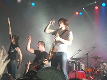 A rock band on stage wearing black or white t-shirts and jeans, in the spotlight performing before an audience.