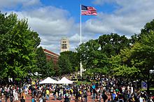 Red brick plaza, surrounded by trees with green leaves, with two white tents and an American flag flying from a flagpole in the center