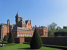 An irregularly shaped red-brick house with slate roofs with an orangery on the ground floor and gables, chimneys and a clock tower above. In the foreground is a formal garden with a lawn, clipped shrubs, a wall and a Victorian-style lamppost.