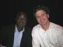 A young black man in glasses and a suit (Valentino Achak Deng) sits to the right of a Caucasian man with wavy black hair wearing a white shirt (Dave Eggers)