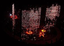 A darkened stage seen from above. Curtains of red and white lights hang down behind and to the sides of the four band members who are lit up with spotlights. Three of the members are on the main stage while the fourth is on the b-stage, an elliptical catwalk that surrounds the main stage.