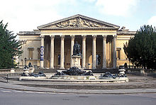 A Palladian style nineteenth century stone building with a large colonnaded porch. In front a large metal statue on a pedestal and fountains with decorations.