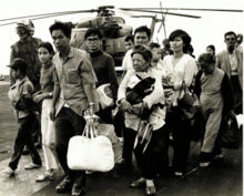 Twelve refugees of varying ages, carrying bundles of possessions, arrive on the deck of a United States naval vessel. Three US airmen, as well as a helicopter, are visible in the background.