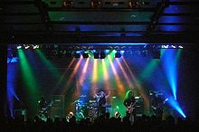 Members of a band, long-haired and dressed in black, perform under multi-colored lights.