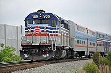 Blue and silver train engine with red and white accent lines moves closer leading a series of similarly colored passenger cars with shrubs and a sound wall in the background.