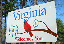 A large square metal sign, mostly white, with the words Virginia Welcomes You in blue and red. In the center a red cardinal bird sits on a branch with two white flowers around it.