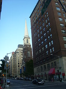 One side of a city street, viewed from an intersection. There is a large brick building on the right. To its left is a tall brick church with a white steeple.