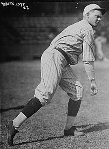 A man wearing a pinstriped baseball uniform stands truned to his left having just thrown a baseball.