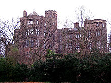 Seen from a low angle through the branches of trees is a substantial stone house with two-three storeys, bay windows and some castellation.