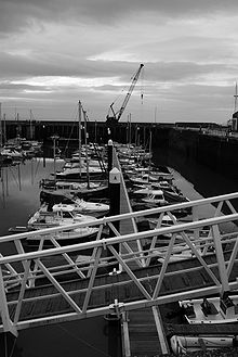small boats lined up in harbour. Crane in the background & metal walkway in the foreground.