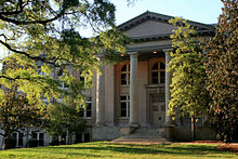 Gray stone building with large Doric columns and grassy foreground, framed by trees
