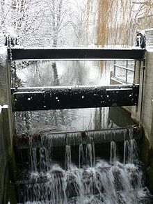 Weir in place of the upper doors of the lock.  This weir has a levelling mechanism attached, rather like a tilting weir. We see the dark frames of the weir, wet on a winter's day, with the water cascading over toward us in a smooth cataract, no turbulence or distrubance.  The surface of the mill pond beyond is placid.  The air is misty and the surrounding trees are white with hoar-frost, which also rimes the edges of the frame of the weir, melted only where the water has splashed.