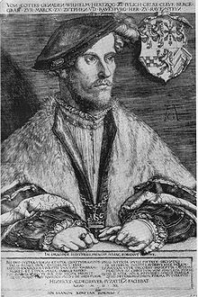 A  middle aged man, Wilhelm, Duke of Jülich-Cleves-Berg, is sitting at a table. He is  dressed in a soft cap that falls to the side of his head. He is wearing  fur-trimmed  robes, and nestled in the edges of the robes is a chain, with a cross  at its nadir. His hands are folded on the table before him. He is  wearing several rings, and one hand holds a pair of gloves. The family  crest hangs on the chair behind him.
