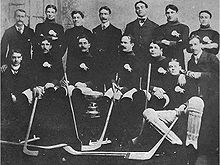 Fourteen men pose around a silver trophy. Several are wearing identical sweaters with a buffalo logo over the left breast and are holding hockey sticks.
