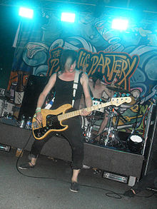 A man wearing a blank tank top, pants that end right before his ankles and black slip-ons plays the bass guitar on a stage. His hair is colored black with a streak of blond over his face and two star tattoos can be seen near his collar bone just under the straps of his tanktop. Behind him a man plays on his drum kit with both his arms raised. The entire stage is layered with different types of carpeting for sound arrangement and a line of blue strobe lamps can be viewed atop of the roof of the stage.