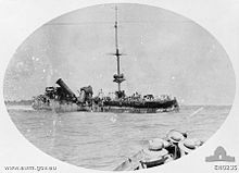A badly damaged ship sits in the water, while in the foreground sailors in another vessel watch on