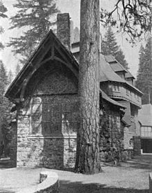 A monochrome photograph of the exterior of a building made of river rock, showing tall stained glass windows at one end, next to the massive trunk of a tall pine tree.
