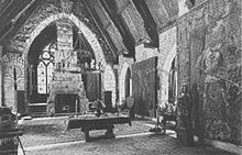 A monochrome photograph of a grand living room with a 36-foot-high arched ceiling, a high fireplace, and tall stained glass windows.