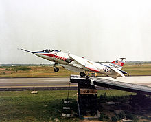 Jet aircraft, with cream and red paint scheme and an American flag on the tail, executing a ski-jump take-off, where an aircraft uses an angled ramp to increase lift before taking off.