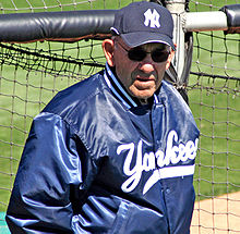 A man wearing sunglasses, along with a navy blue jacket and baseball cap that have white lettering.
