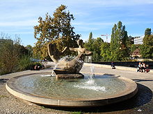 Circular fountain surrounded by pavement. The fountain's center is a sculpture of a pair of abstract human figures.
