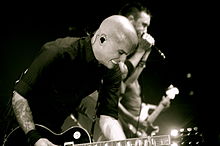 A man with a shaved head playing a guitar with an intense expression. A harmonica player is in the background.