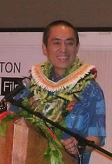 A middle-aged Chinese man standing at a podium, wearing a Hawaiian shirt and lei
