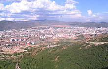 Aerial view of a city, surrounded mostly by desert. In the foreground and background are hills with sparse vegetation.