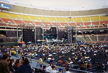 An elaborate concert stage, seen during the day inside a mostly empty stadium. The stage comprises several dark, rectangular structures. Fans are scattered throughout the floor seats, while the stadium seating is empty.