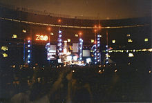 An elaborate concert stage set bearing a logo that reads "Zoo TV", set in a dark stadium. Towers reach into the nighttime sky, illuminated in blue with red warning lights on top.