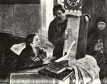 A boy is sitting at a table and is looking at a large open book held by a bearded standing man. The table is covered with books and papers. A woman is sitting by the wall and looking at the man
