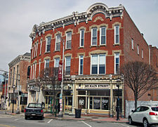 Two joined three-story brick buildings that curve away from the camera to their right, seen from across the street. They have ornate rooflines that rise to semicircles in the middle, elaborate stone lintels over their windows and ornate wooden storefronts at street level. The one on the right has "145 Main Street" in large letters above its window