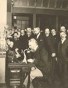 A grizzly and well-dressed Alexander Graham Bell sits at a desk talking over antique telephone, surrounded by numerous business executives and news reporters, who are witnessing a historic event, in the atrium of a large corporate building.