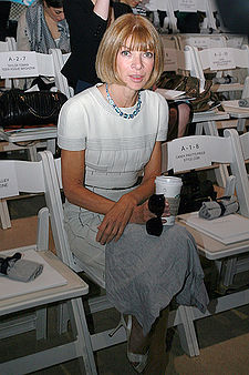 A seated woman wearing a white dress, holding a coffee cup and sunglasses, looking at the camera. The surrounding seats are empty.