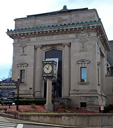A small square stone building with ornate decorative work, including the words "Bank for Savings". In front of it is a small clock tower with "Time to enjoy Historic Ossining" on it. At lower left is a sign welcoming visitors to the Ossining Central Business District.
