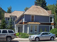 Another side of the same house seen in the first picture. This side is longer, with only two windows on the first story and one above, topped by a peaked roof of light-colored wood. Two silvery cars are parked on the street in front.