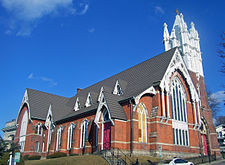  A brick church, seen from a corner and looking uphill, with a tall white steeple. One of its front windows has been boarded up.