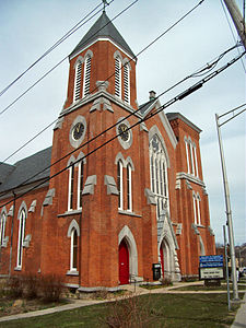 A brick building with narrow windows and vents seen from its right, its upper sections partially obscured by telephone wires. Closest to the camera is a tower with a pyramidal gray top and clocks on both visible faces showing 11:05