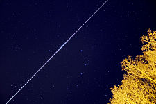 A view of a dark blue, starry sky with a white line visible from the bottom-left to the top-right of the image. A tree is visible to the bottom right.