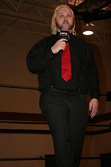 A male wrestler dressed casually in a wrestling ring holding a microphone