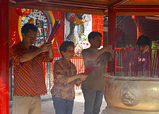 A middle-aged man, an elderly woman, and two younger men pray in front of a censer vessel while holding red incense sticks. They are in a roofed structure painted in red.