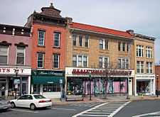 Three brick buildings seen from across the street, with a portion of a fourth visible on the left. The one next to it is the tallest, with "Keenan" in large block letters on its brown pediment and "1878" visible below. The next building is the widest, made of yellow brick with a red tiled roof sloping steeply downward. Its storefront is taken up by a "Healthsmart Pharmacy". The building on the right is also of yellow brick, occupied by "Lillian's Youth Center"